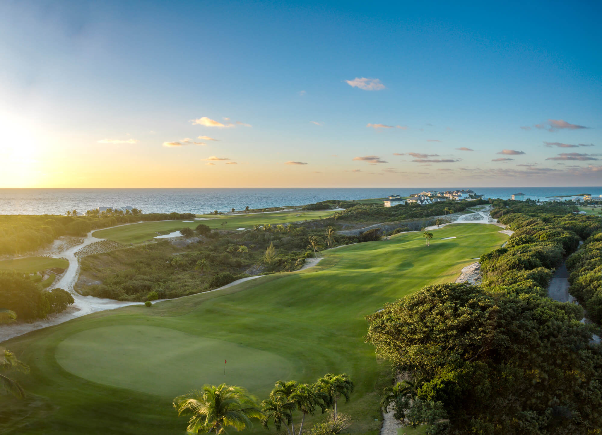 Sunset over a coastal golf course with green fairways, sand traps, and palm trees near the ocean horizon.