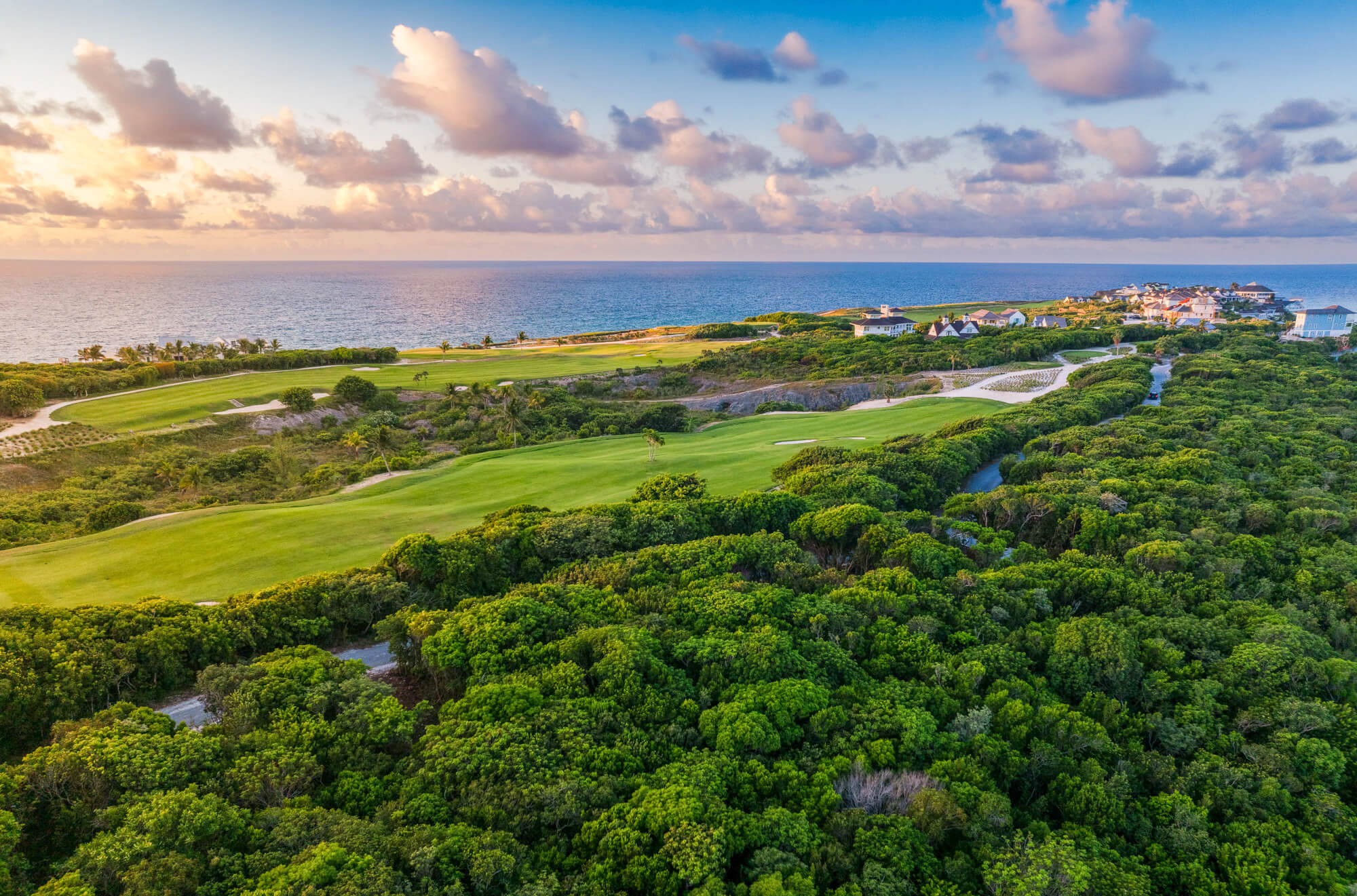 Aerial view of a coastal golf course with green fairways, blue ocean, and houses along the shore on the right.
